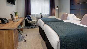 A hotel room with a wooden desk, two beds, a chair, a TV, and a window with curtains.
