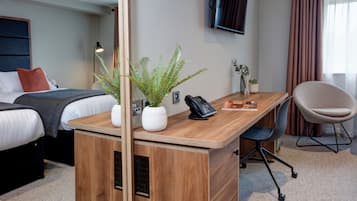 A hotel room with a bed, a desk with a phone, a chair, a TV, and a plant.