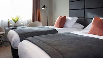 A hotel room with two beds, a bedside table with a lamp, a potted plant, and a view of the outdoors.