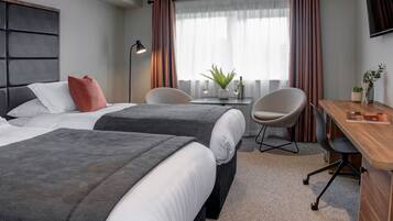 A hotel room with two beds, a desk, a chair, a small table, and a plant.