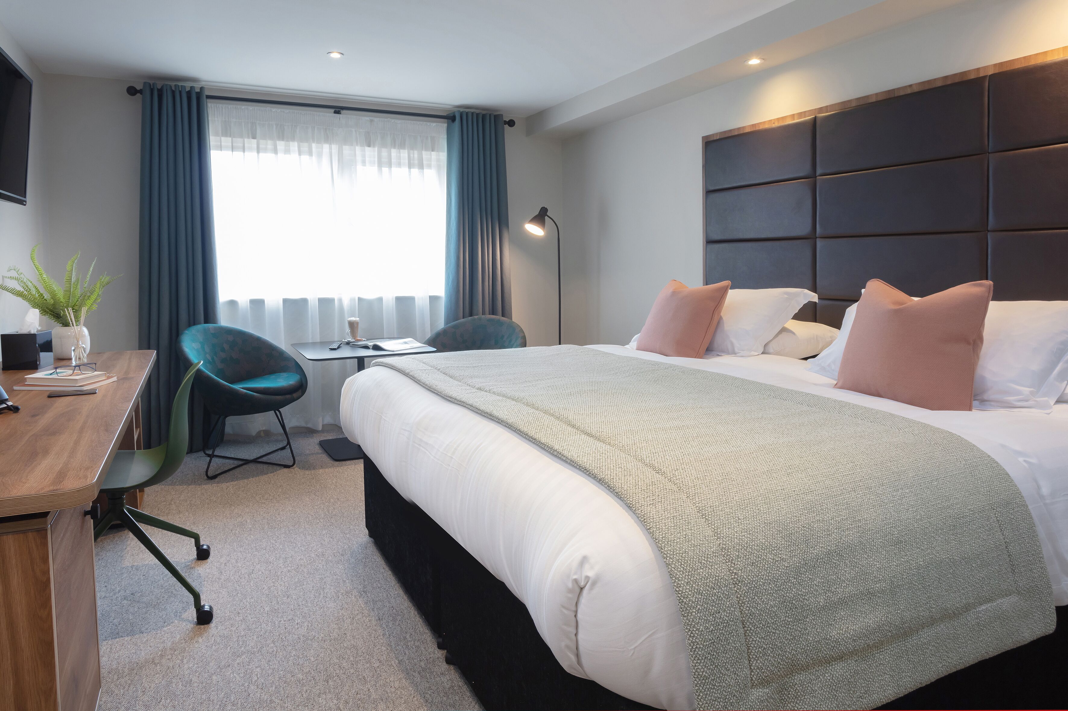 Quarto de hotel com uma cama grande, uma mesa, uma cadeira e uma janela com cortinas.