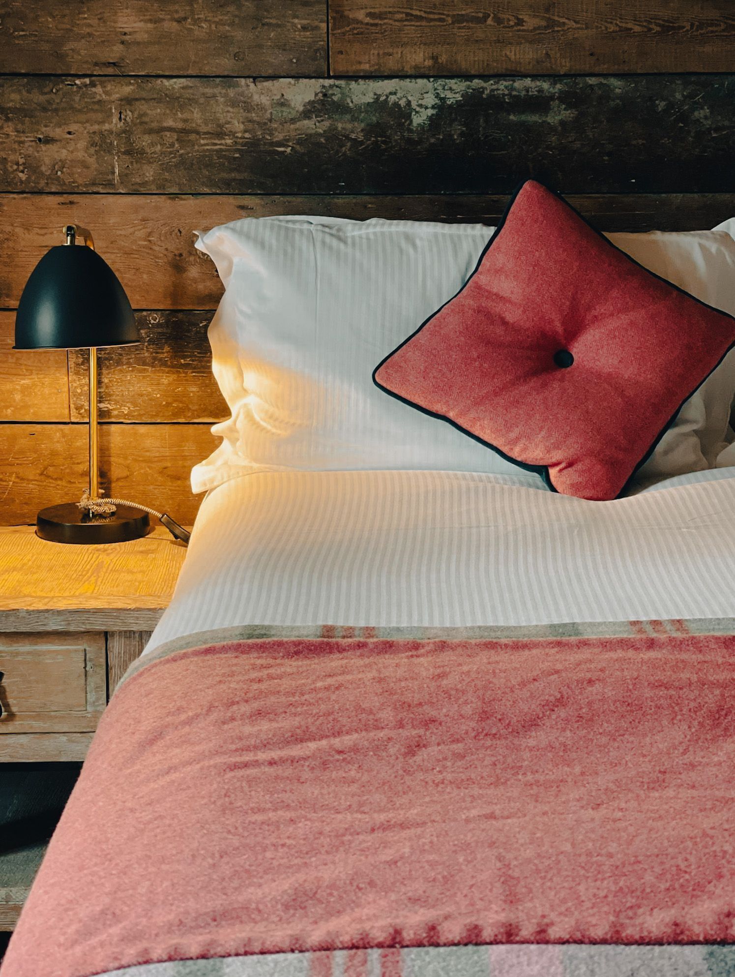A bed with a white comforter and a pink blanket, a red pillow, and a black lamp on a wooden nightstand.