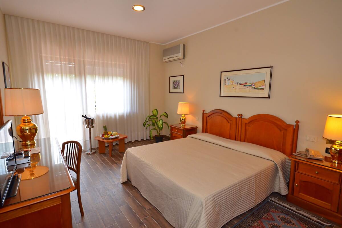 Une chambre d’hôtel avec un lit, des tables de chevet, un bureau avec une lampe, une chaise, une plante en pot et un tableau au mur.