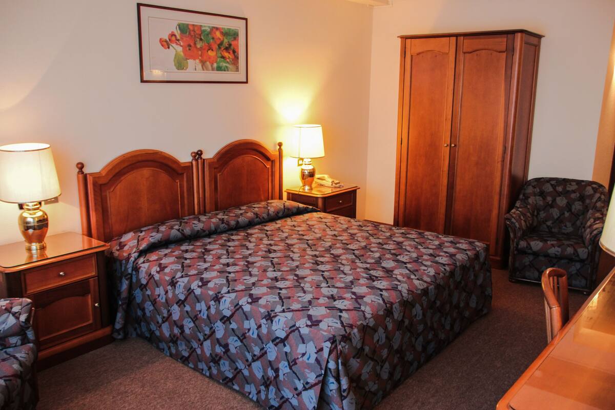 Une chambre d’hôtel avec un lit en bois, deux tables de chevet avec des lampes, une armoire, un fauteuil à motifs et une œuvre d’art florale encadrée accrochée au mur.