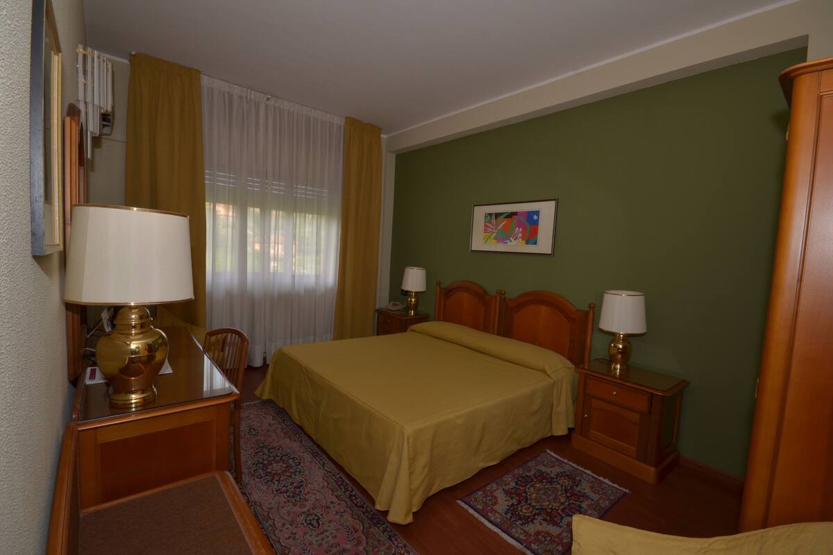 Une chambre d’hôtel comprenant un lit, deux tables de chevet avec des lampes, une chaise, un bureau avec une lampe, une fenêtre avec des rideaux et un tableau au mur.