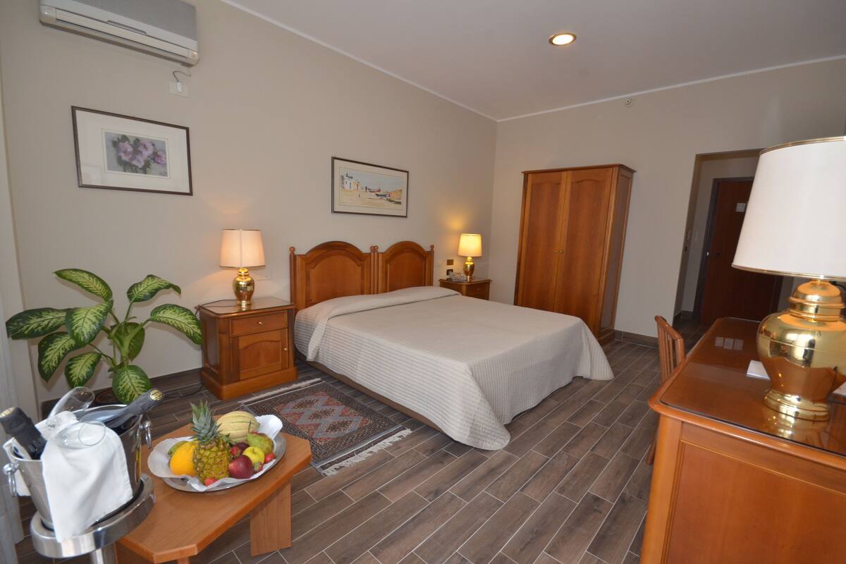 Une chambre d’hôtel avec un lit, des lampes de chevet, une armoire en bois, un bureau avec une lampe, un panier de fruits et une plante en pot.