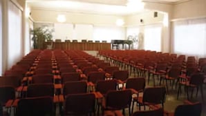 A conference room with rows of chairs and tables, large windows, and a stage area. - Garden Hotel (San Giovanni la Punta)