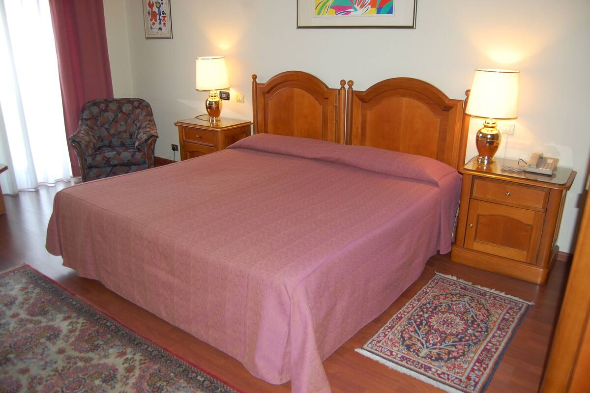 Un lit bien fait, recouvert d’une couvre-lit rose, deux tables de chevet avec des lampes, un fauteuil à motifs floraux et un tapis à dessins.