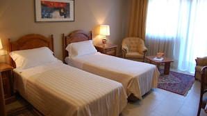 A hotel room with two beds, a chair, a small table, a lamp, and a painting on the wall. - Garden Hotel (San Giovanni la Punta)