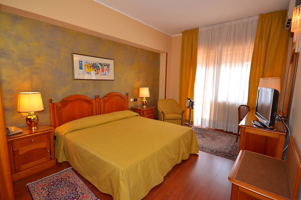 Une chambre d’hôtel avec un lit, deux tables de chevet, une chaise, un téléviseur et un tableau au mur.