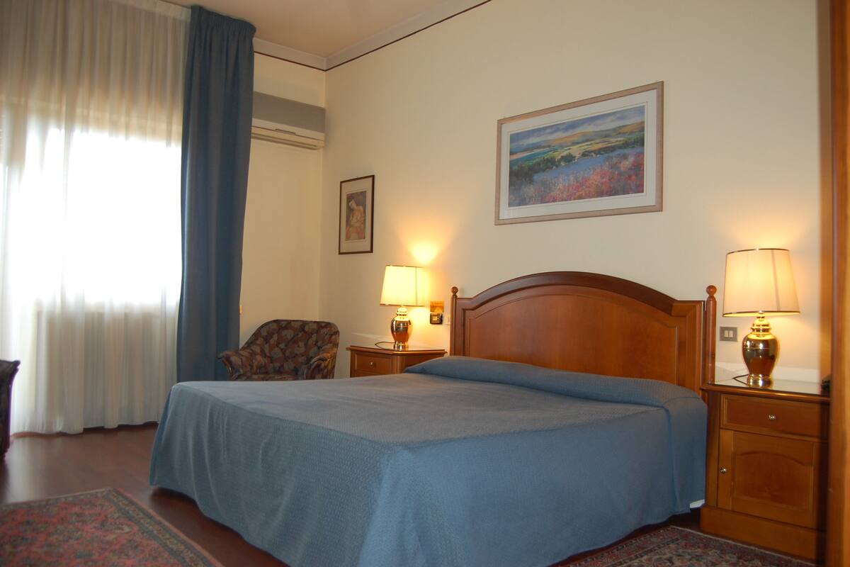 Une chambre d’hôtel avec un lit, deux lampes de chevet, une chaise, une table de chevet, un tableau au mur et une fenêtre avec des rideaux.