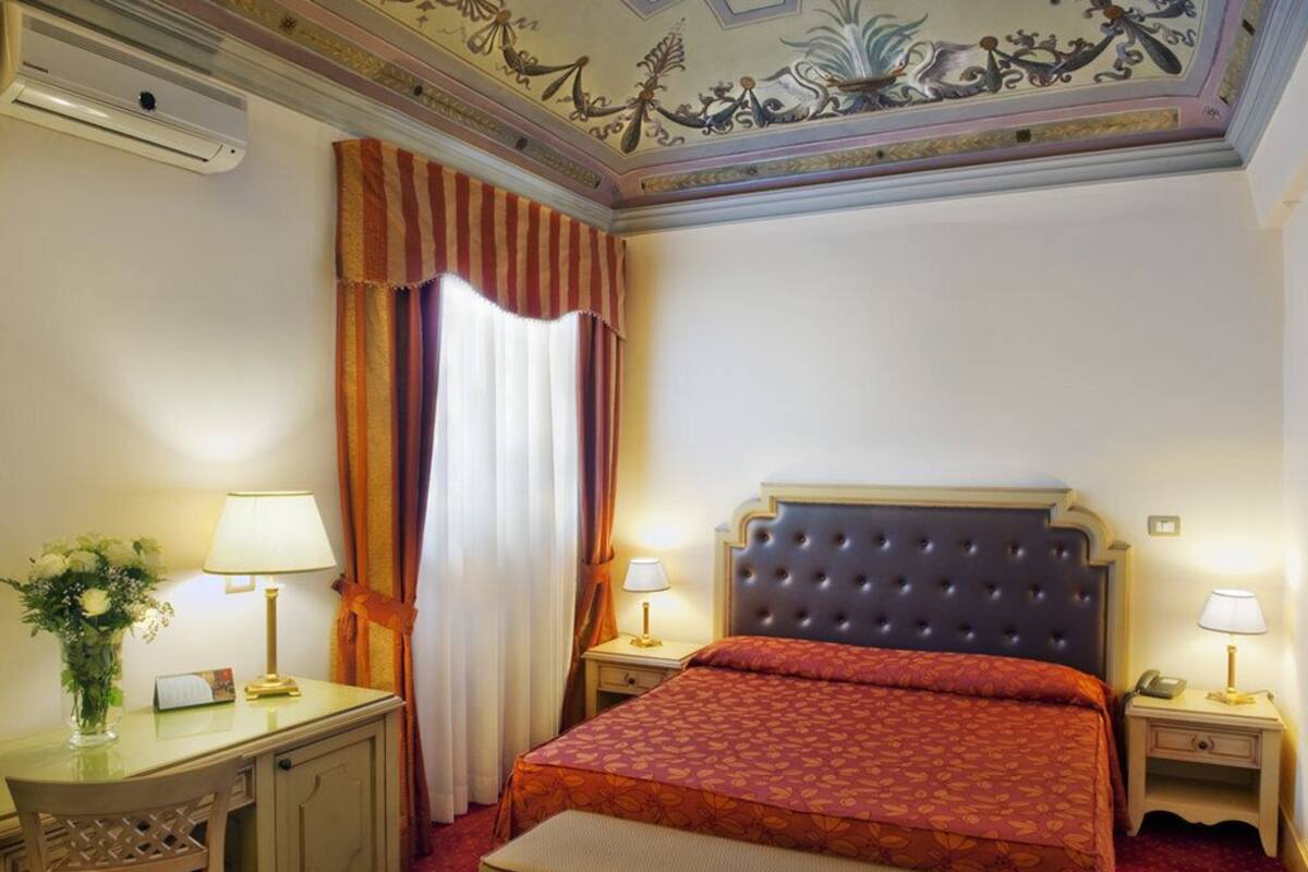 Habitación de hotel con cama, dos mesitas de noche con lámparas, un escritorio con lámpara, una silla y un techo decorativo.