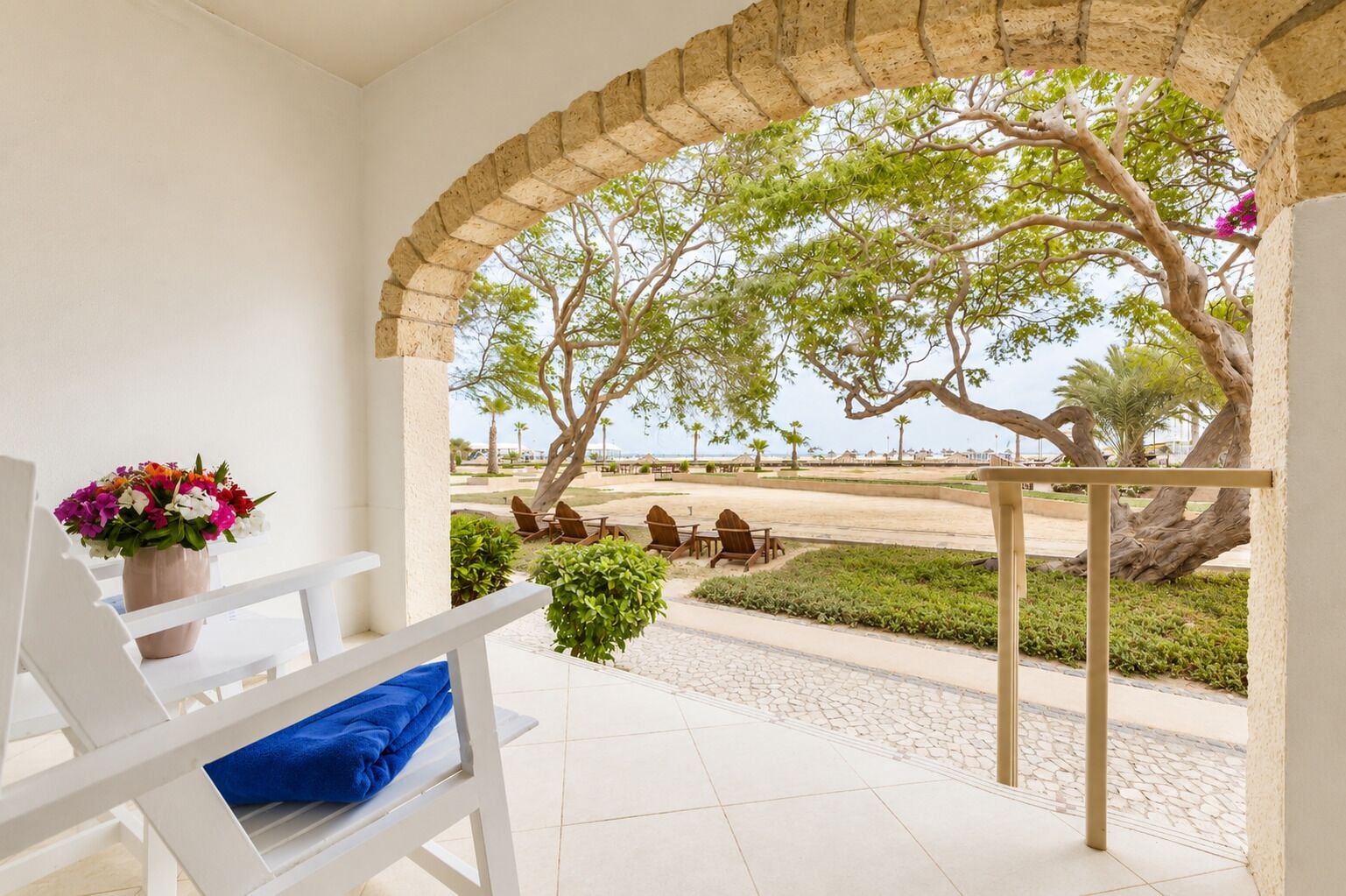 Un balcon donnant sur une plage, une chaise avec un coussin bleu et un vase de fleurs.