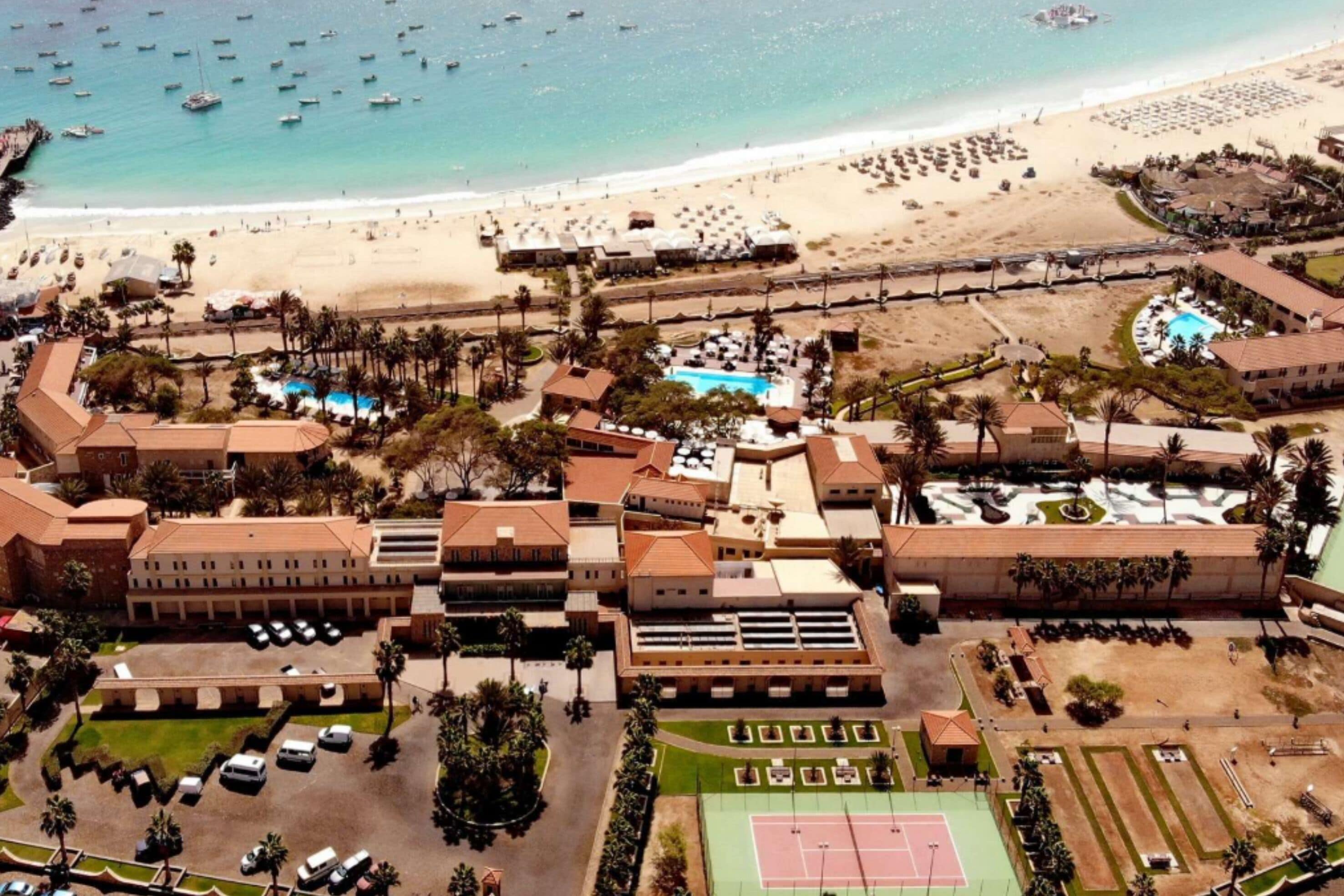 An aerial view of a resort with a beach, swimming pool, tennis court, and multiple buildings.