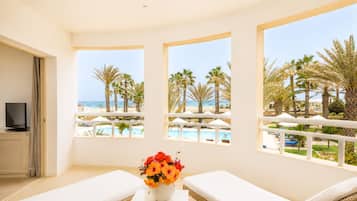 Een kamer met balkon, witte meubels, een televisie en uitzicht op strand en zwembad.