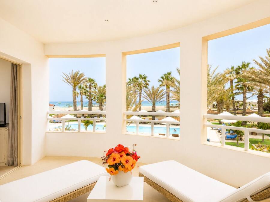 A room with a balcony, white furniture, a TV, and a view of a beach and pool.