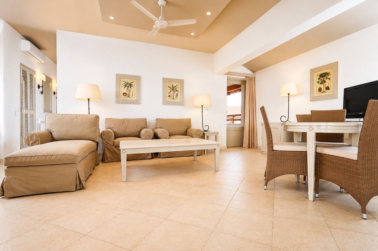 A spacious living room with a sofa set, a dining table, and chairs. There is a ceiling fan, wall-mounted lamps, and framed pictures on the walls.