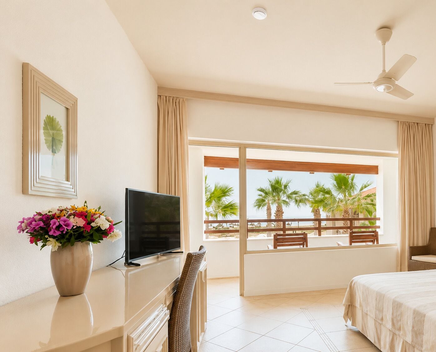 Habitación de hotel con televisor, florero, cama, balcón y ventilador de techo, con vistas a palmeras.