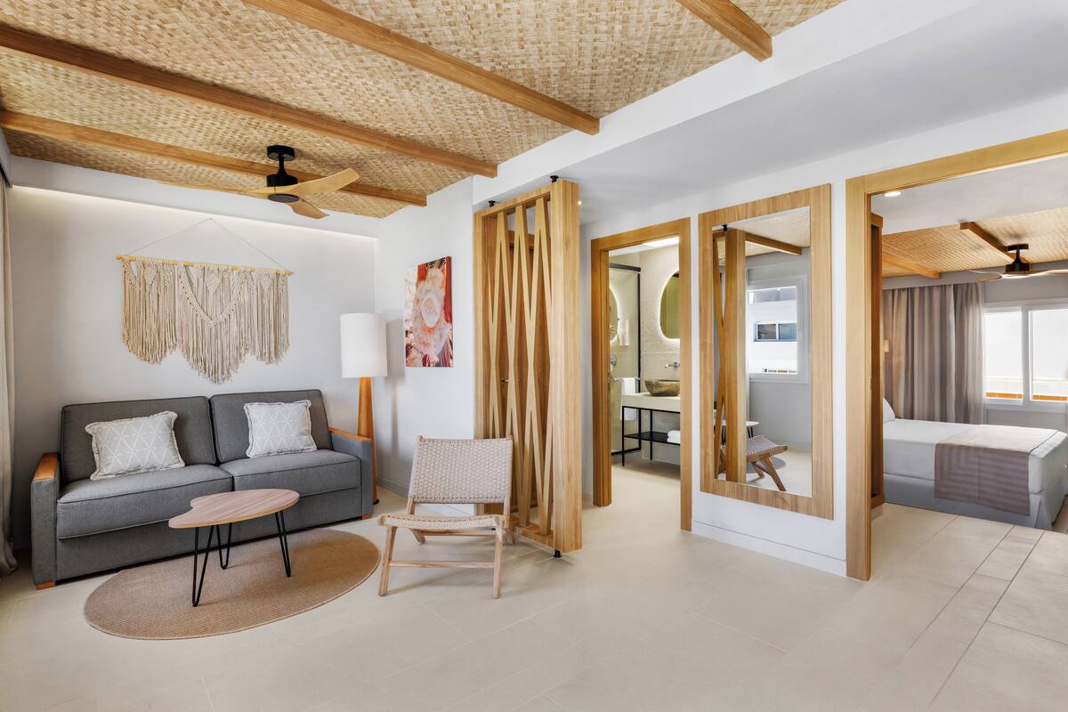 A modern living room with a grey sofa, a wooden coffee table, a woven chair, and a large mirror.