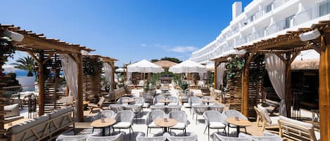 An outdoor seating area with wicker chairs and tables, wooden pergolas, and umbrellas.