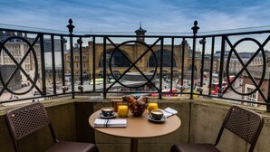Ein Balkon mit Frühstückstisch, gedeckt mit Kaffee, Saft und Croissants, mit Blick auf eine belebte Straße und historische Gebäude.