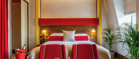 A hotel room with a bed, a small table with two glasses of champagne, a red chair, and a large window.