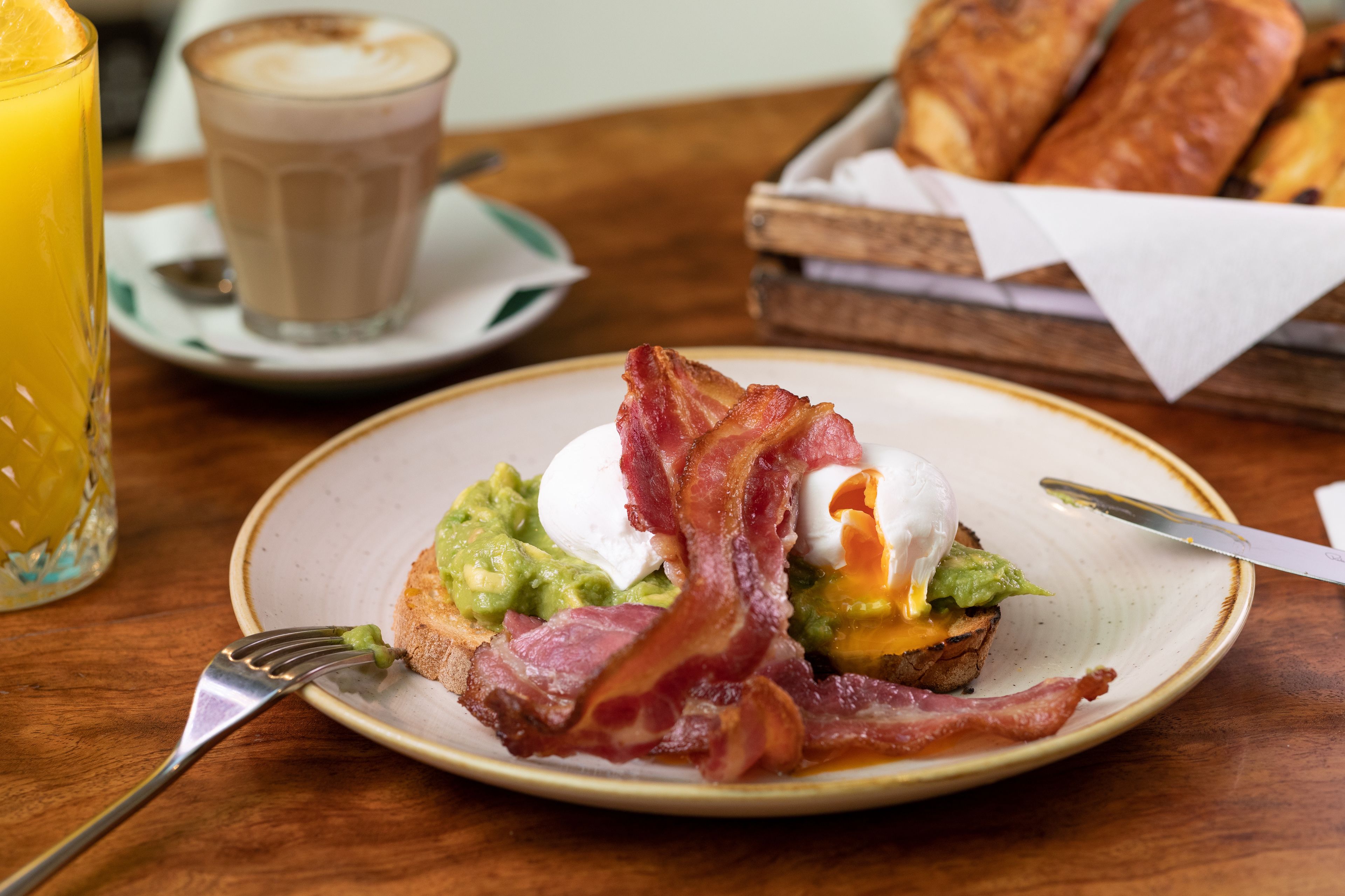 Een ontbijtje met avocado, ei en bacon op toast, een glas sinaasappelsap en een kop koffie op een houten tafel.