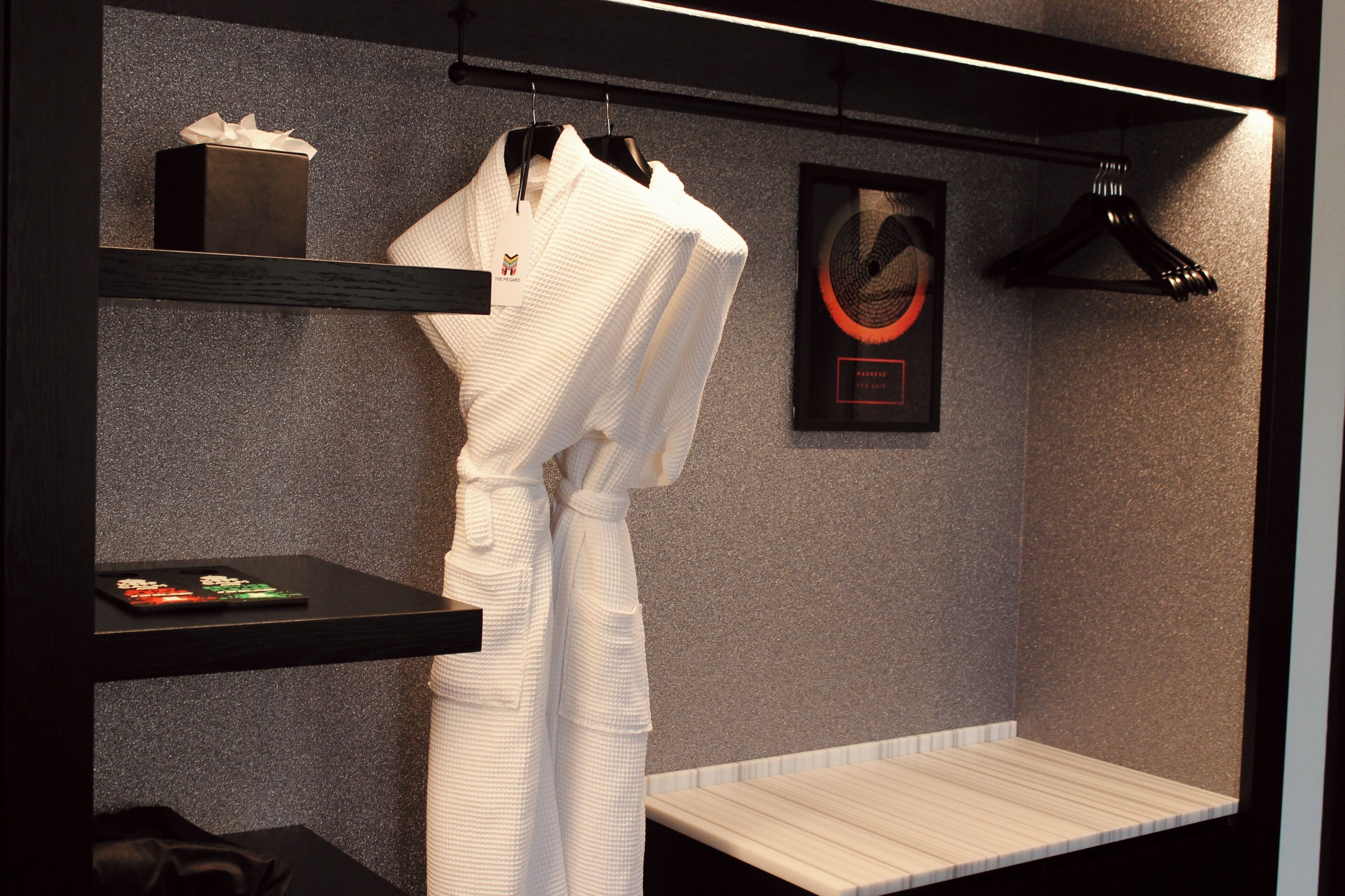 A spa room with a wooden bench, a white robe hanging on a rack, and a framed artwork on the wall.