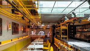 Ein Restaurant mit hohen Decken, gelb-weißer Einrichtung und einer Bar mit Regalen voller Flaschen.