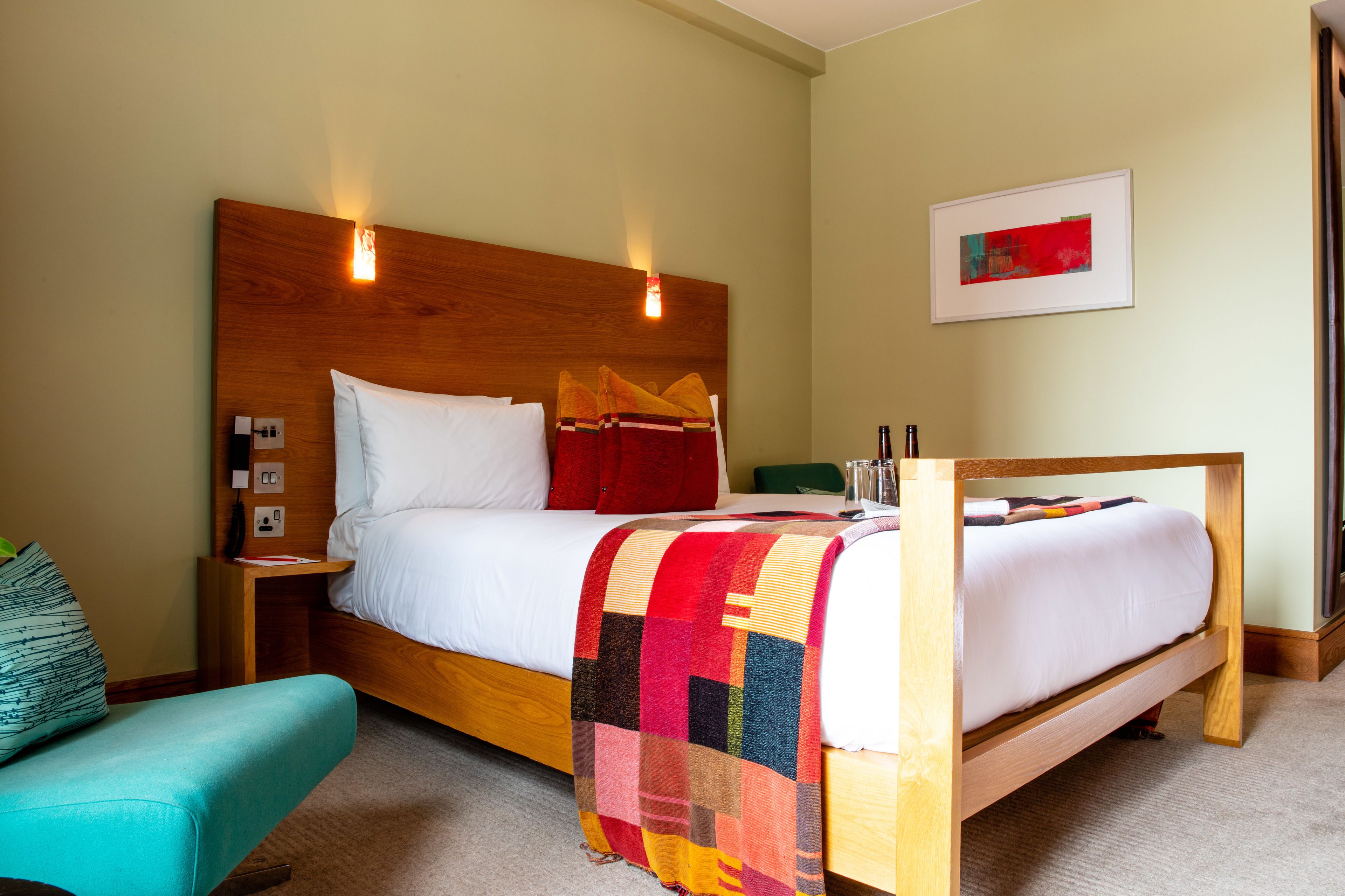 A hotel room with a wooden bed, a colorful blanket, a teal armchair, and a framed picture on the wall.