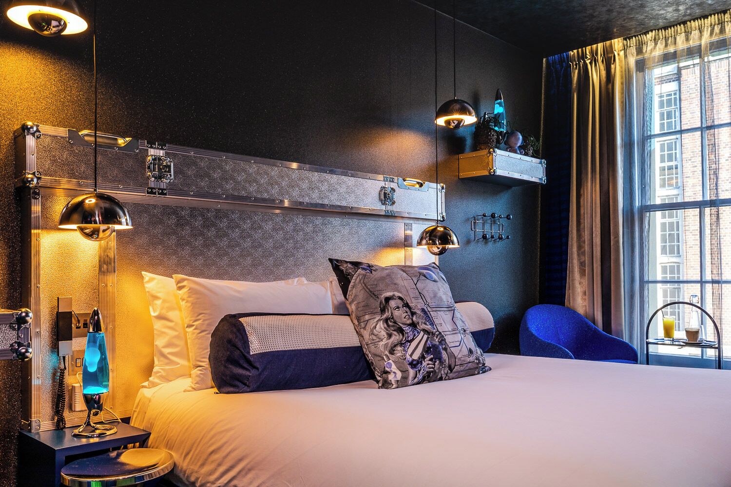 A modern hotel room with a large bed, a nightstand with a lamp, a blue chair, and a window with curtains.