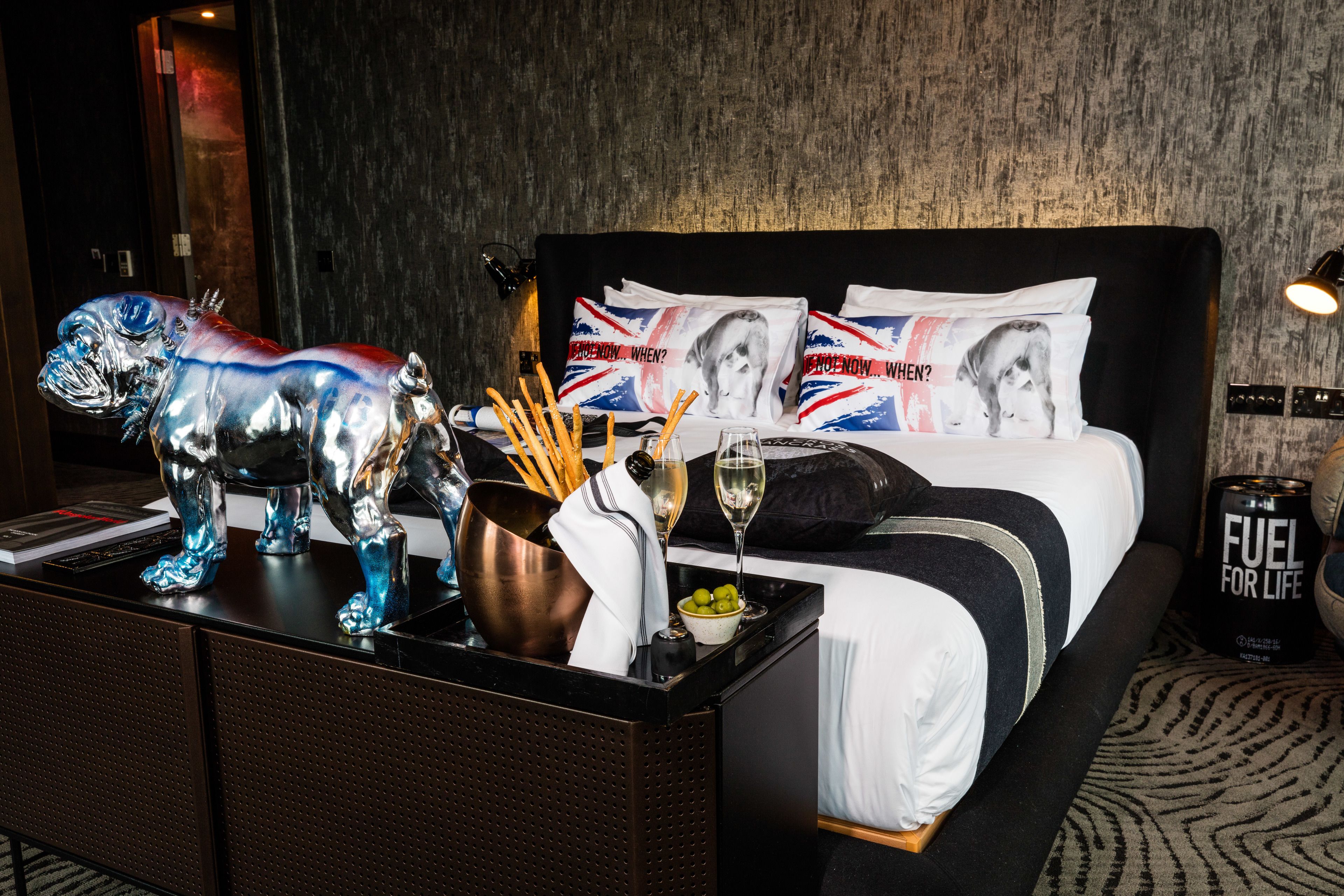Habitación de hotel con cama, mesita de noche, escultura metálica decorativa, un cubo, una copa de vino y una lata de combustible.