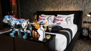 Habitación de hotel con cama, mesita de noche, escultura metálica decorativa, un cubo, una copa de vino y una lata de combustible.