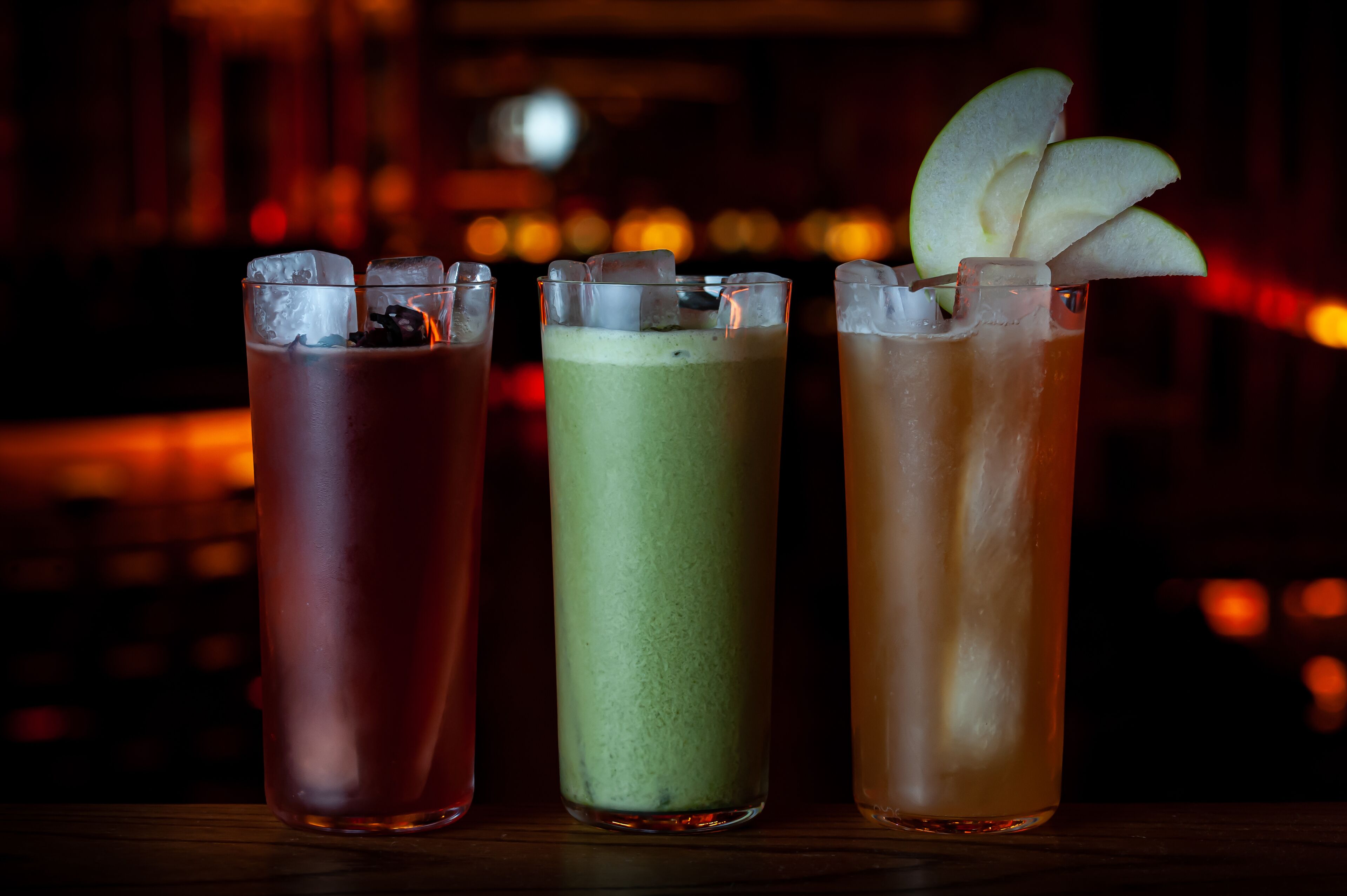 Tres vasos de bebidas de colores diferentes con cubitos de hielo y adornos sobre una barra.
