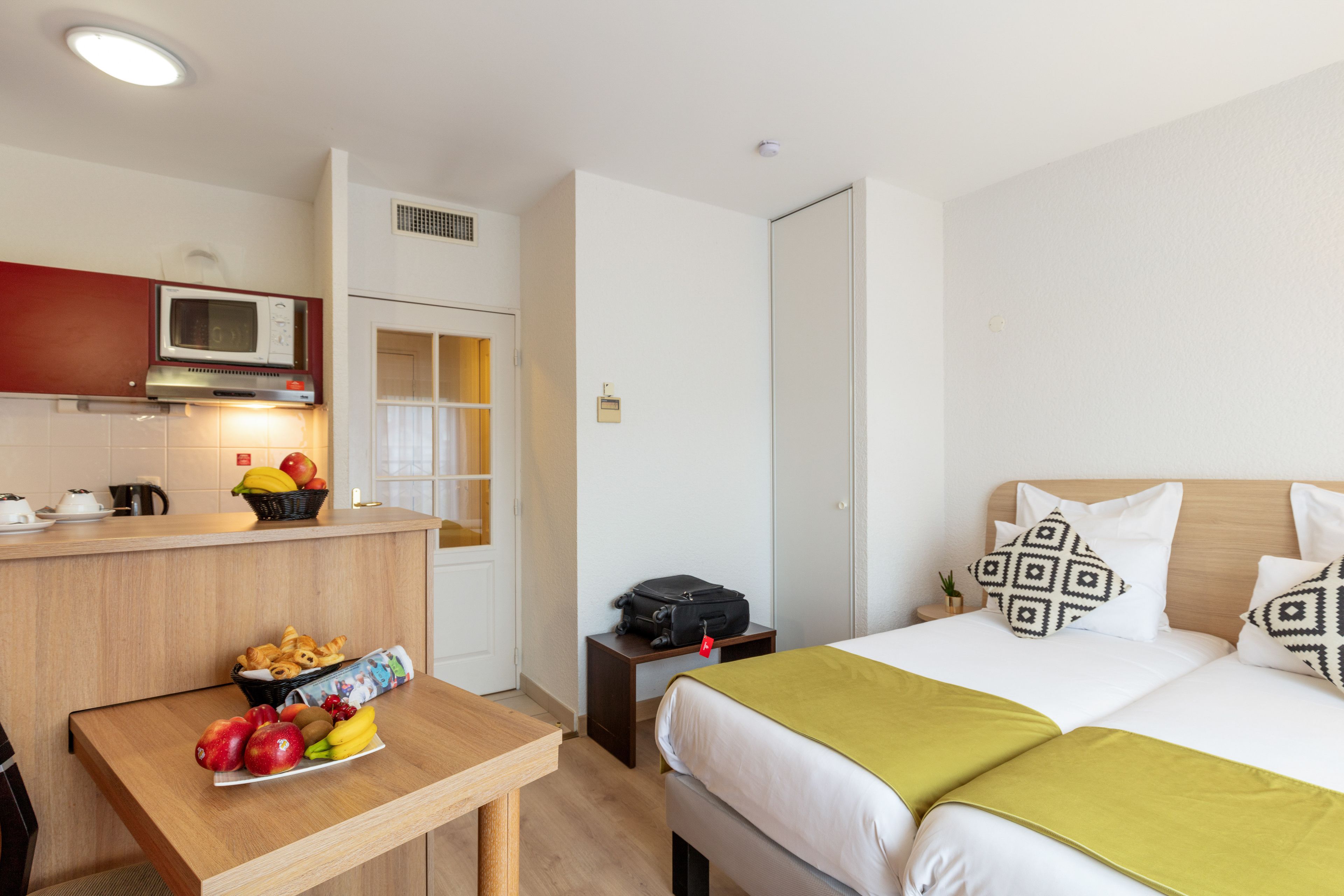 Habitación de hotel con cama, mesita de noche con frutas, microondas y kitchenette pequeña.