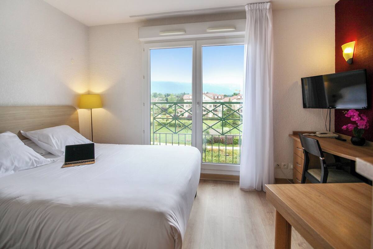 Habitación de hotel con cama, escritorio con silla, televisor y balcón con vistas a edificios.
