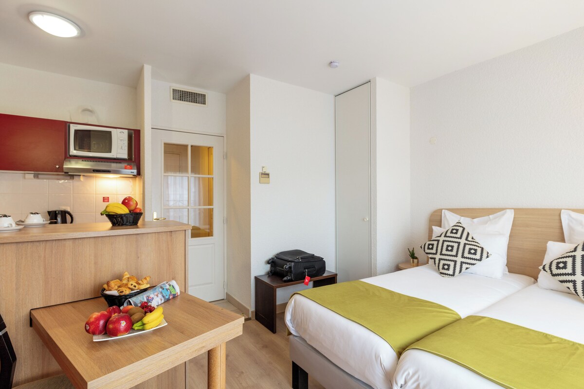 A hotel room with a bed, a bedside table with fruits, a microwave, and a small kitchenette.