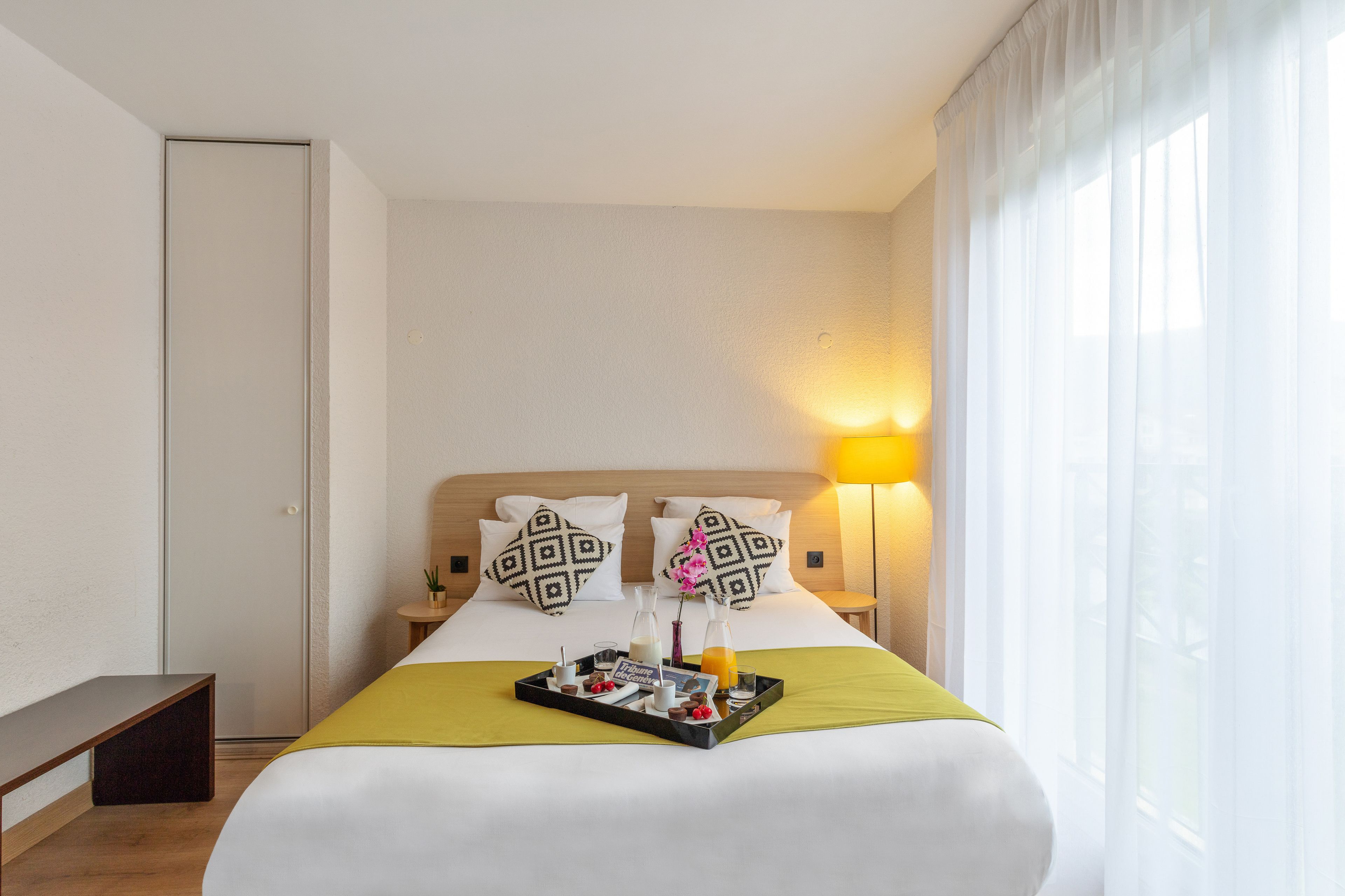 Ein Hotelzimmer mit Bett, Nachttisch, Lampe, Fenster mit Vorhängen und einem Tablett mit Frühstücksartikeln.