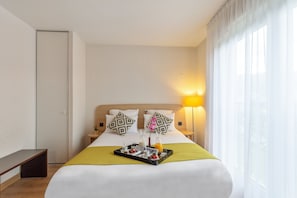 Habitación de hotel con cama, mesita de noche, lámpara, ventana con cortinas y una bandeja con alimentos para el desayuno.