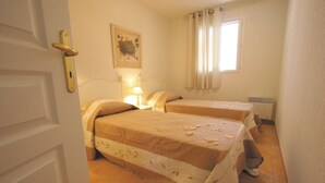 A room with two beds, a nightstand with a lamp, a window with curtains, and a door with a handle.