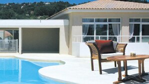 A pool area with a chair and table, a building with a tiled roof, and a view of a hill with trees.