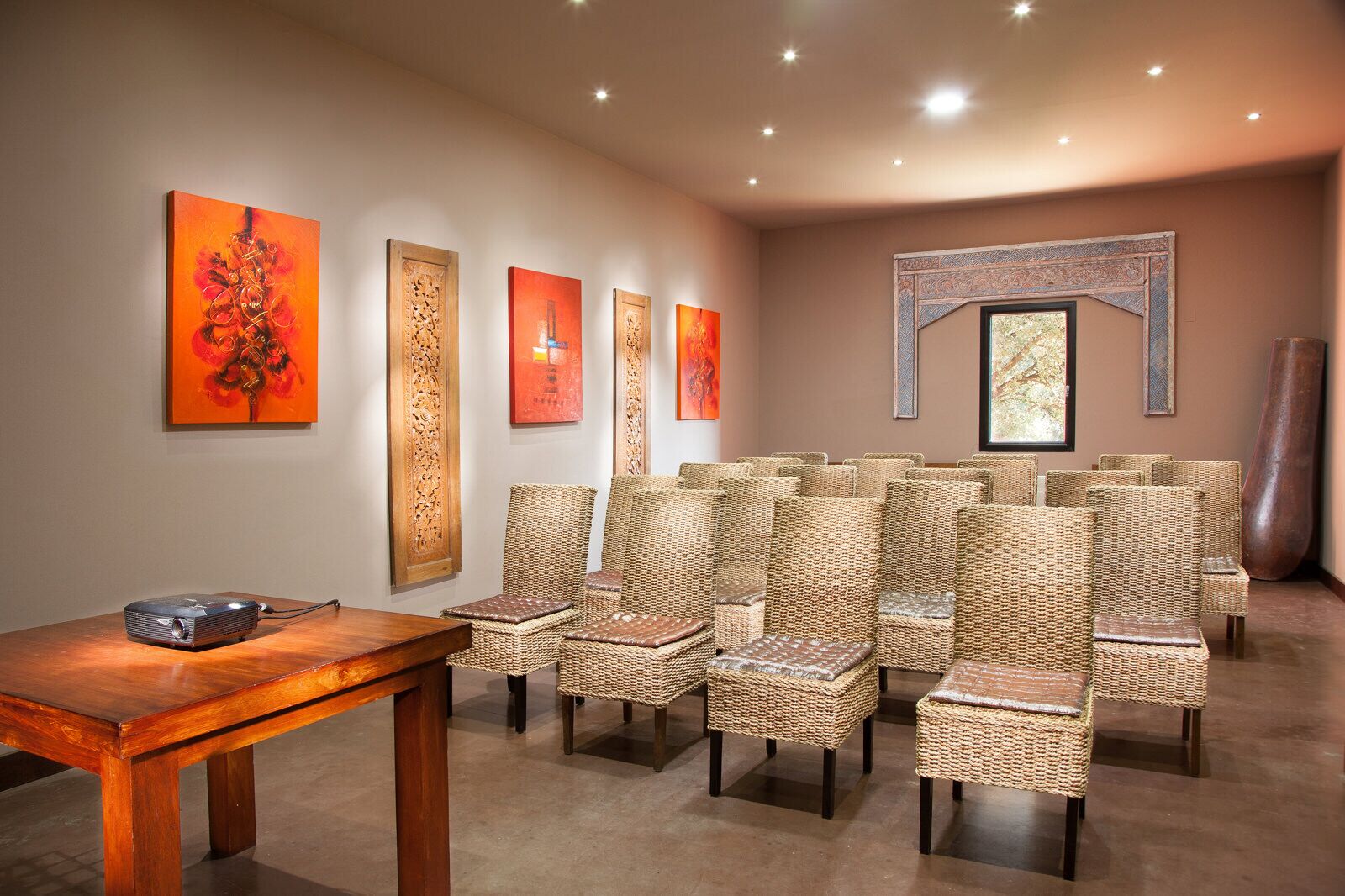 A room with wicker chairs arranged in rows, a wooden table, and artwork on the walls.