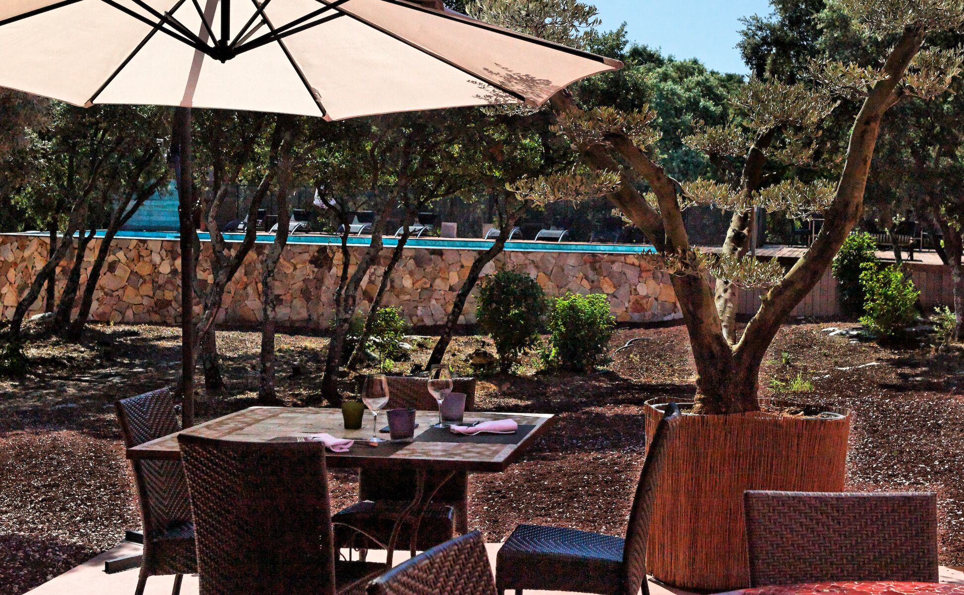 An outdoor dining area with wicker chairs and a table, a large umbrella, and a stone wall with a pool in the background.