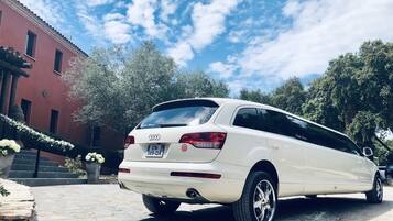 Un coche familiar blanco estacionado en un camino de adoquines, con un edificio rojo y árboles al fondo.
