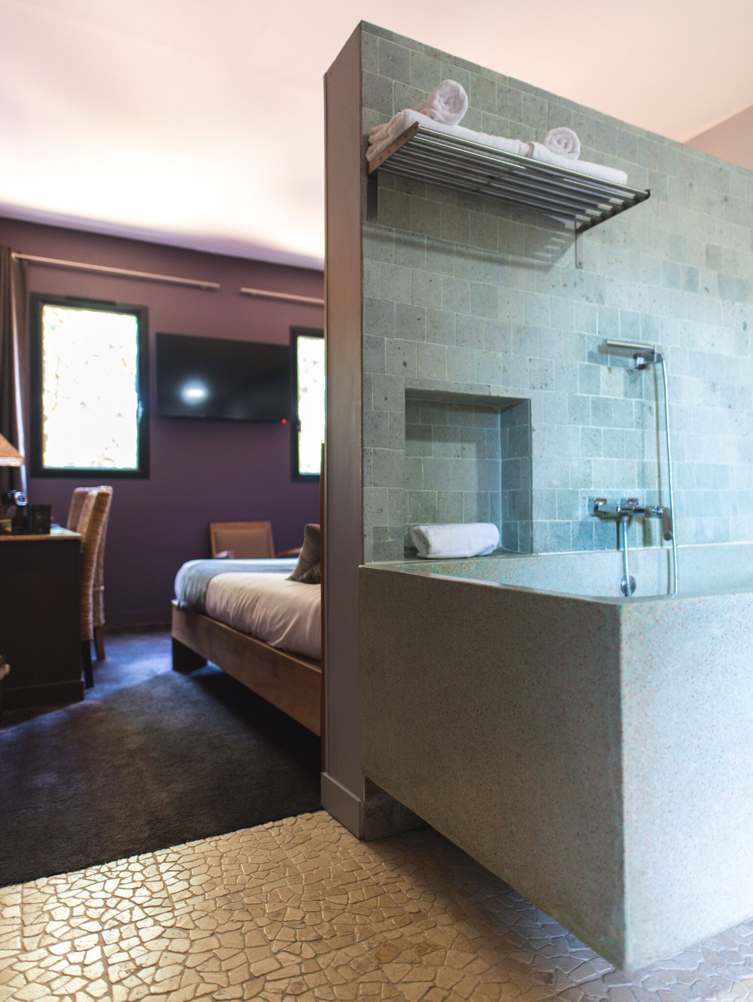 A bathroom with a glass-enclosed shower, a towel rack, and a bed in the adjacent room.