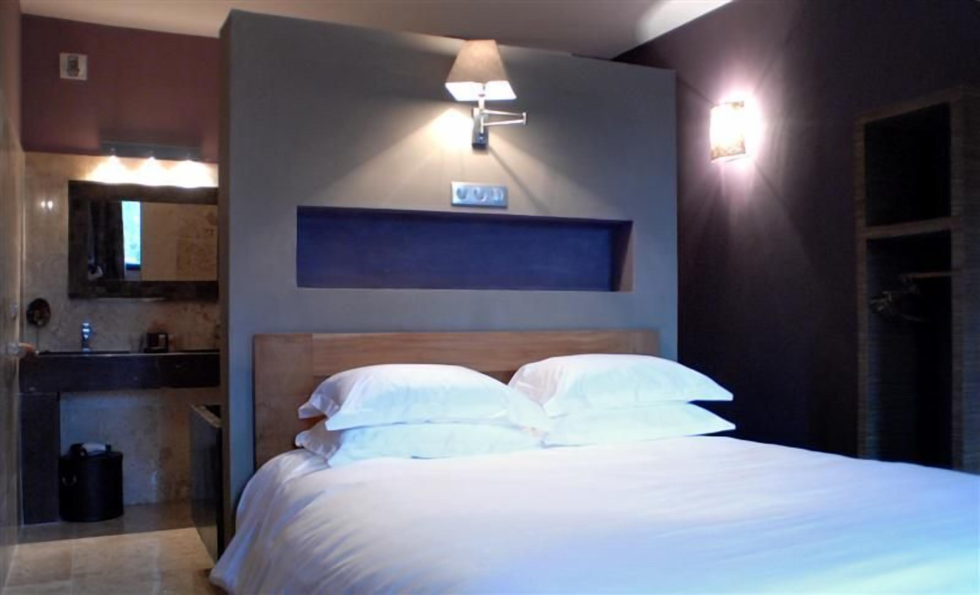 Habitación de hotel con cama, mesita de noche, espejo y lámpara de pared.