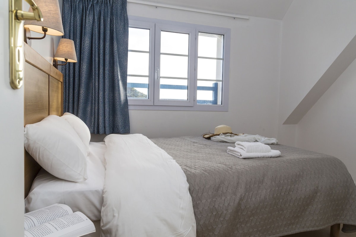 Quarto com cama, almofadas, mesa de cabeceira com abajur, janela com cortinas e toalha na cama.
