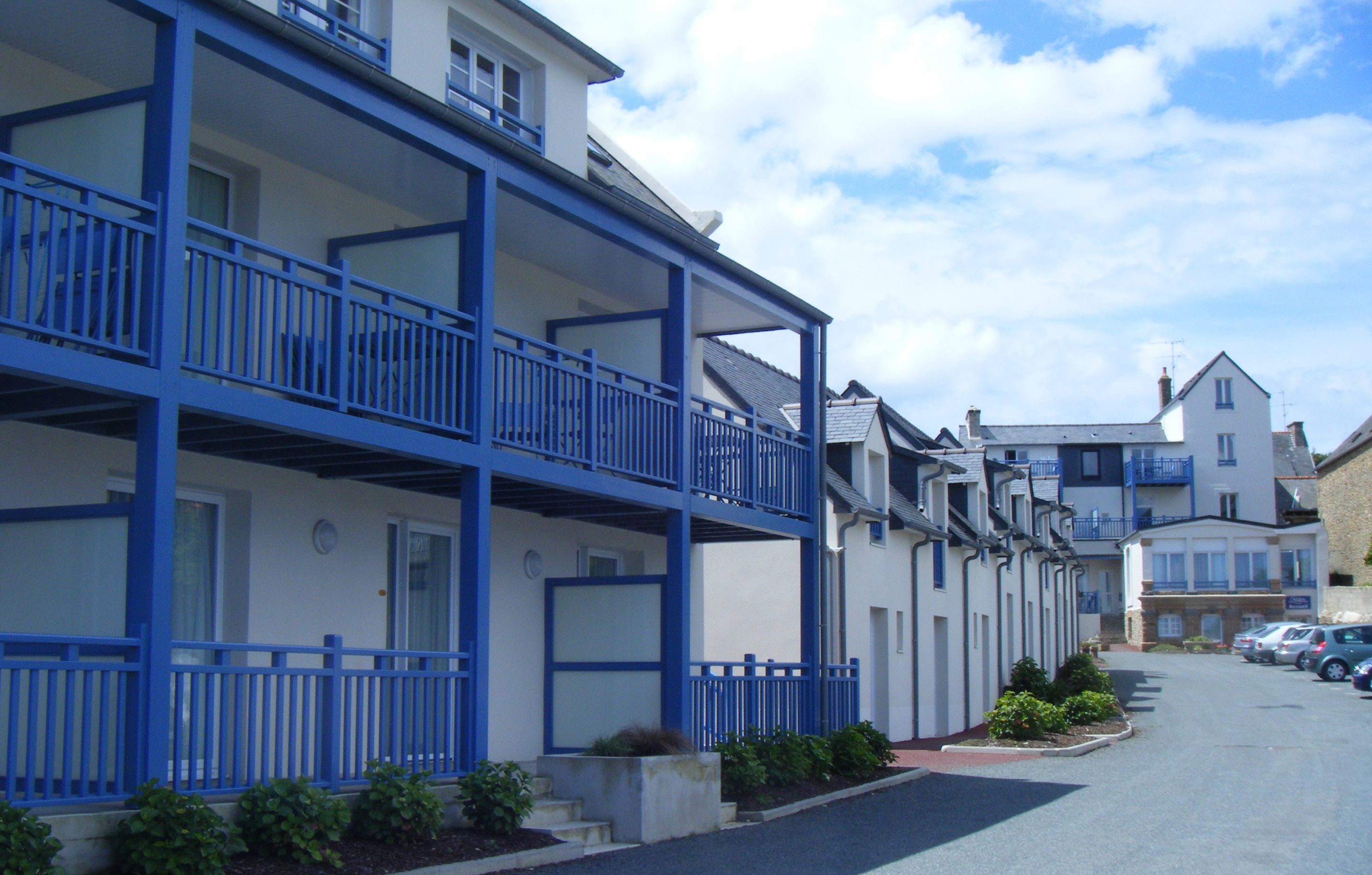 A row of white buildings with blue balconies and railings, a parking lot, and a clear sky.