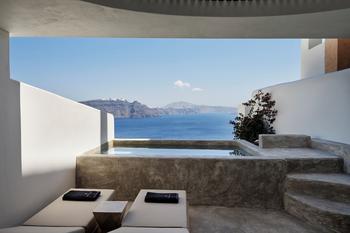A modern outdoor terrace with a pool, two lounge chairs, and a view of the sea and distant cliffs.