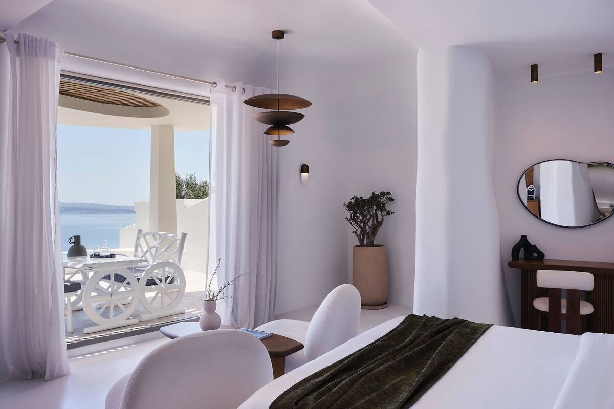 A modern hotel room with a balcony, white furniture, a large potted plant, and a view of the sea.